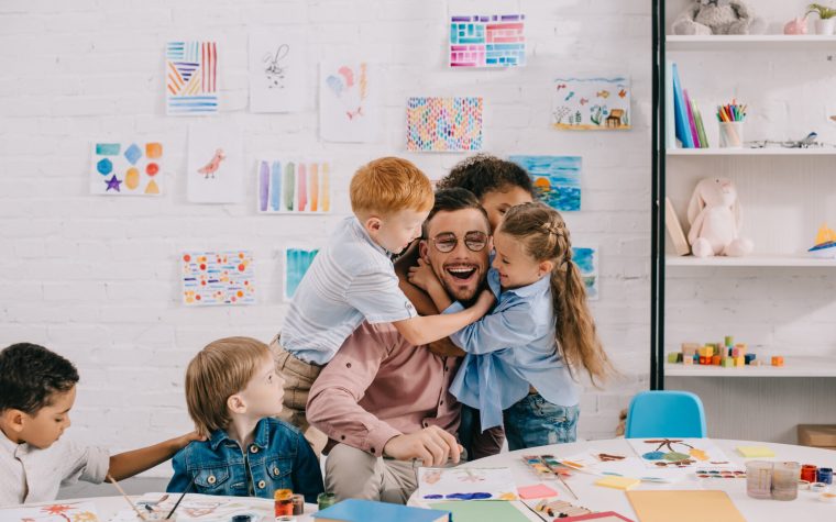 interracial kids hugging happy teacher at table in classroom