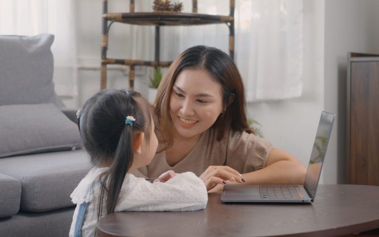 Asian young mother with laptop computer teaching her kid to learn or study online in living room at home, Mum and little preschool daughter learning online on computer, family homeschooling online