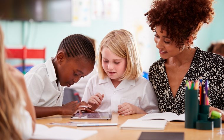 Female Teacher With Two Elementary School Pupils Wearing Uniform Using Digital Tablet At Desk